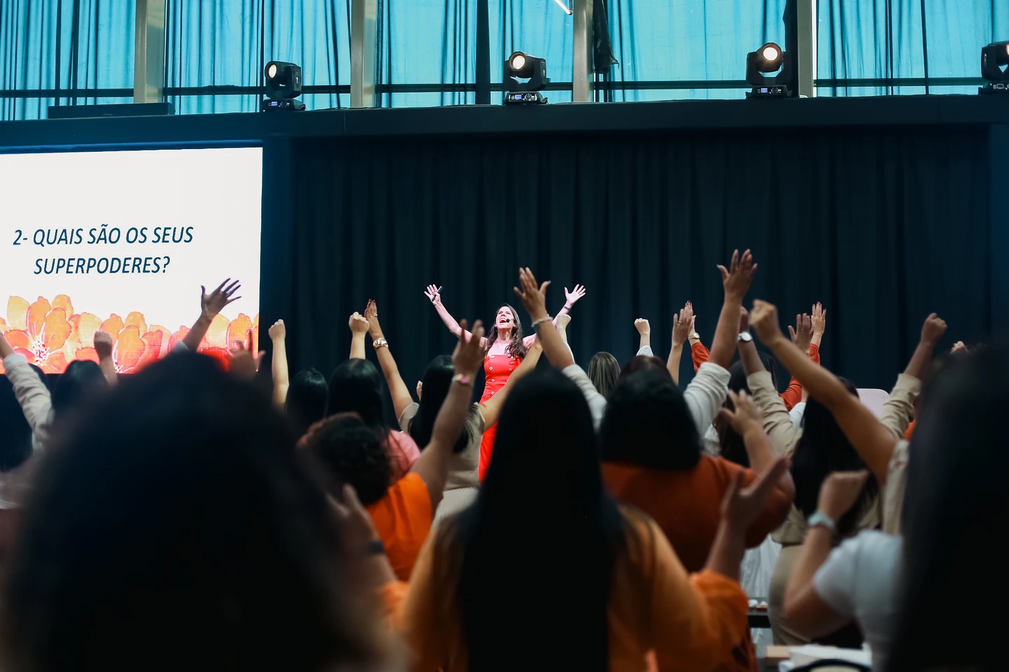 Renata André no palco, conduzindo uma palestra com a participação do público de braços levantados.